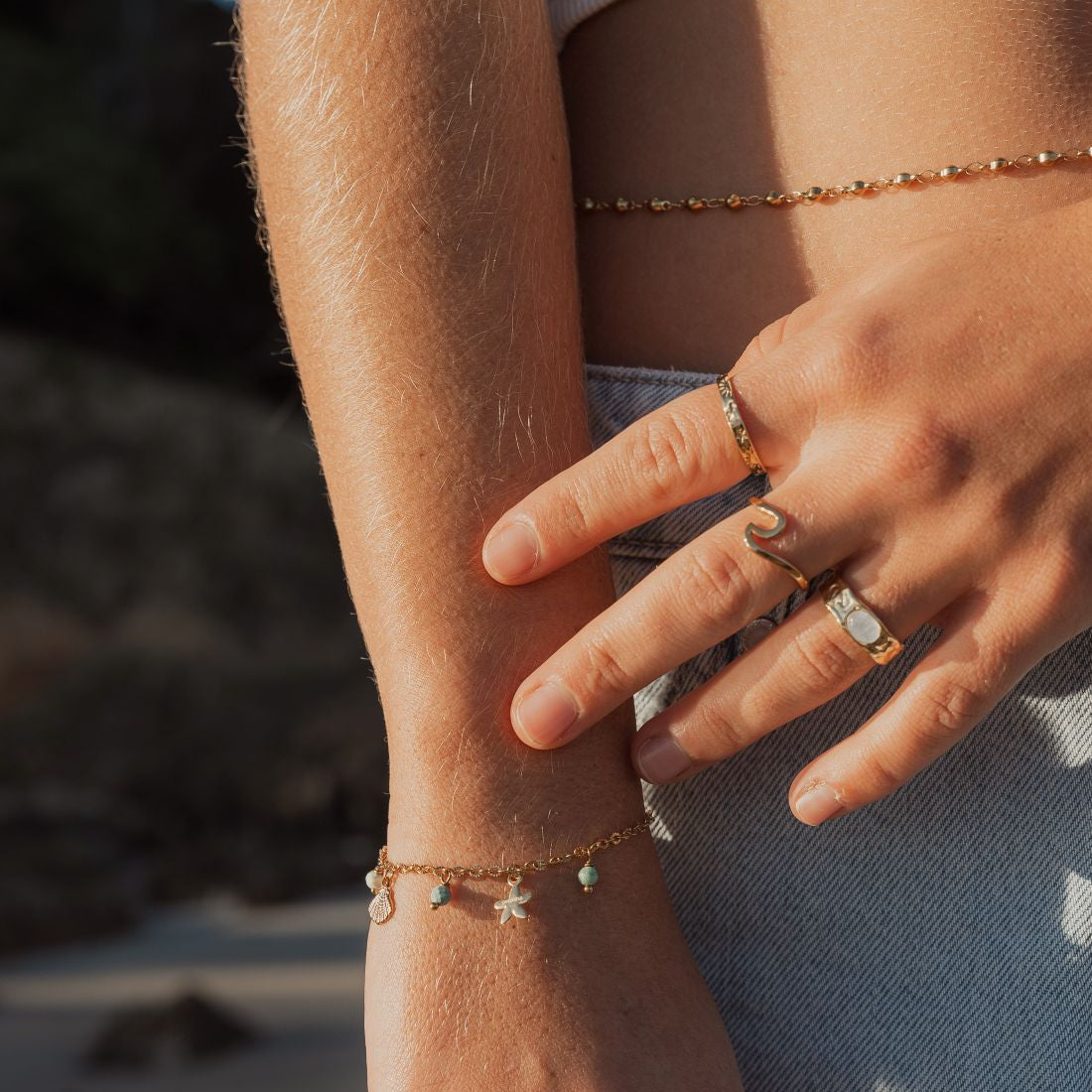A close-up lifestyle shot of a person's hands and arms adorned with multiple delicate gold jewelry pieces, including rings, bracelets, and a chain necklace, set against a softly blurred beach background.