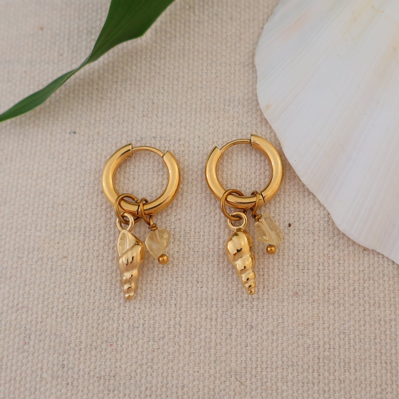 Gold hoop earrings with charms on a beige surface with a shell and leaf.
