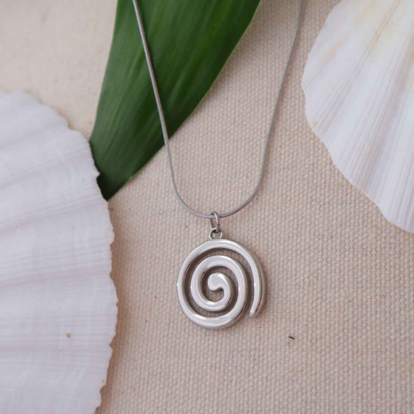 Silver spiral pendant necklace on a beige fabric background with seashells and green leaves.