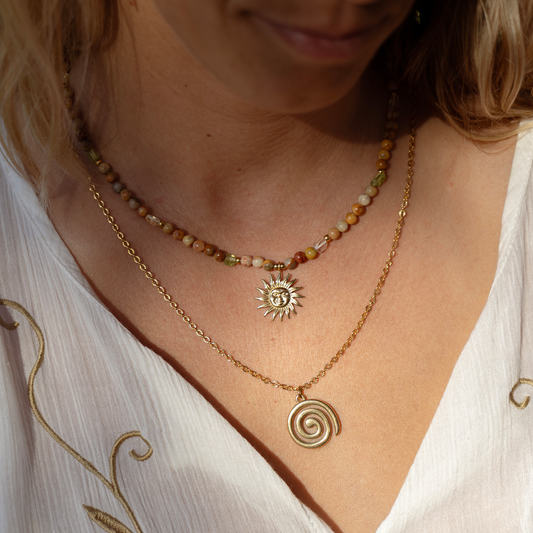 Close-up of a person wearing layered necklaces: a short beaded necklace with earthy multicolor beads, a gold sun pendant, and a longer gold chain with a spiral pendant, paired with a white embroidered top.