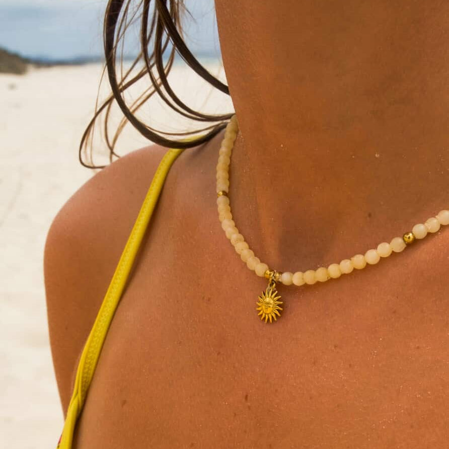 Person wearing a yellow bikini top with a necklace featuring a sun pendant on a beach.