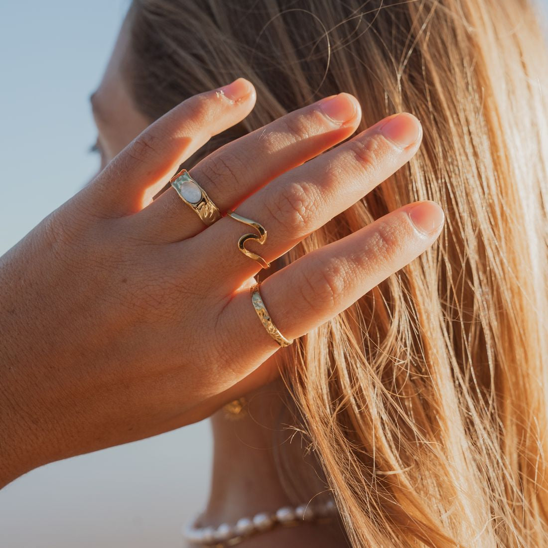 A close-up lifestyle shot of a woman's hand adorned with three gold rings, including a textured band, a wavy ring, and a thin band, set against her long blonde hair and a pearl necklace.